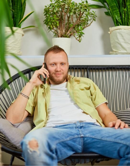 A man in a yellow shirt and ripped jeans sits on a black chair, talking on a phone. There are potted plants behind him.