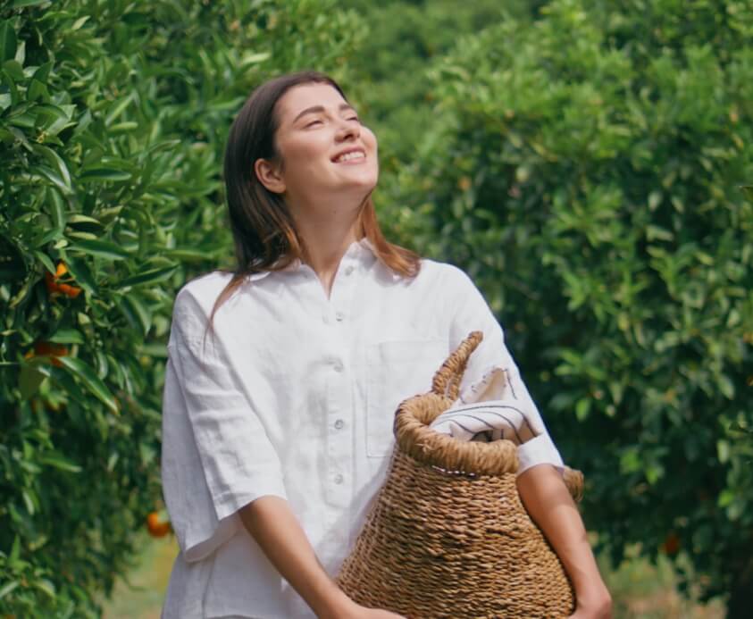 Woman in a white shirt smiling while holding a wicker basket in an orchard.