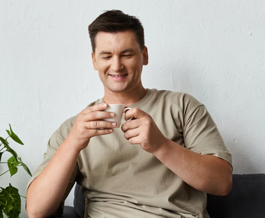 Person in a beige shirt sitting on a couch, smiling at a mug they are holding with both hands. A green plant is next to them.
