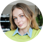 A person with wavy blond hair wears a green sweater over a blue striped shirt, smiling slightly in an indoor setting with a plant and office items in the background.