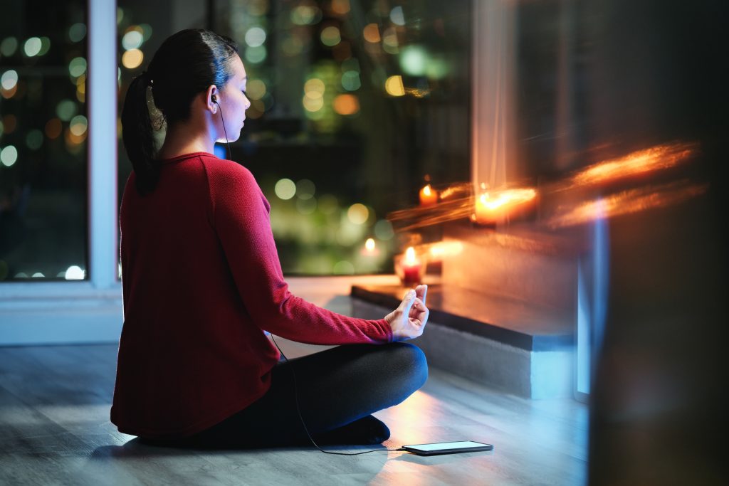 A person sits cross-legged on the floor, facing a window with city lights, practicing meditation near lit candles, seeking relief from sleep problems through serene mindfulness.