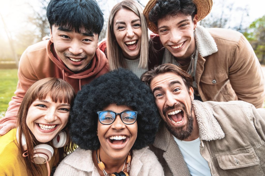 A group of six friends smiling and laughing outdoors, with trees in the background. They are dressed casually and enjoying a sunny day, embracing the joy of friendship while understanding anxiety and promoting mental health.