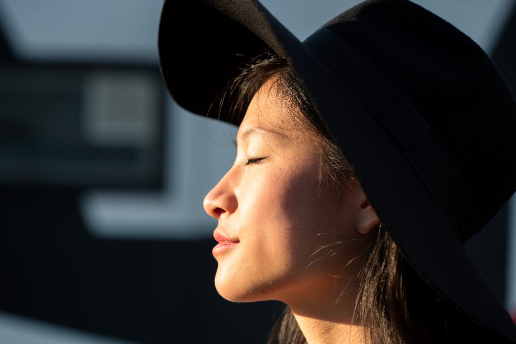 A person with long hair and a wide-brimmed hat enjoys the sunlight, eyes closed, in an outdoor setting, embracing a moment of mood control and feeling better.