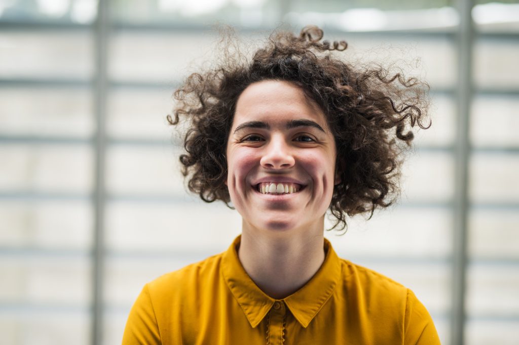A person with curly hair, emanating a cheerful mood in a yellow shirt, is smiling in front of a blurred, grid-patterned background.