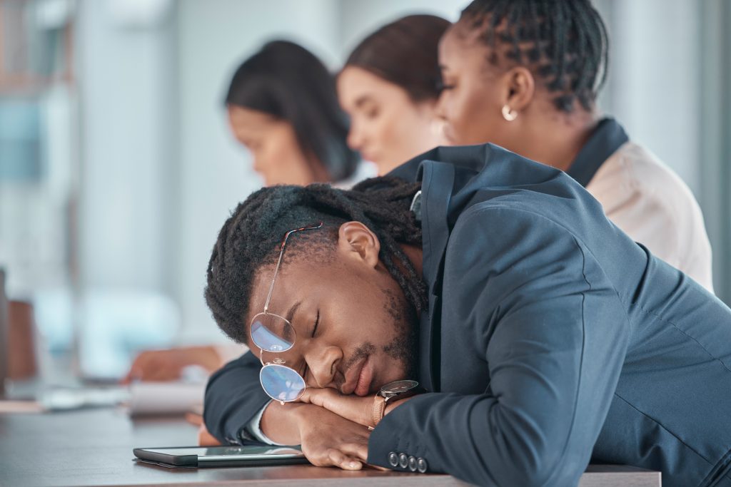 A person in a suit, struggling with sleep problems, dozes at a desk with a smartphone and glasses nearby, while three others work diligently in the background.