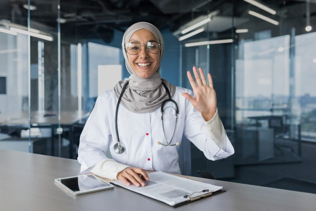 A doctor in a lab coat and stethoscope, radiating a cheerful mood, smiles and waves from behind a desk adorned with documents and a tablet—a practical guide to feeling better right at your fingertips.